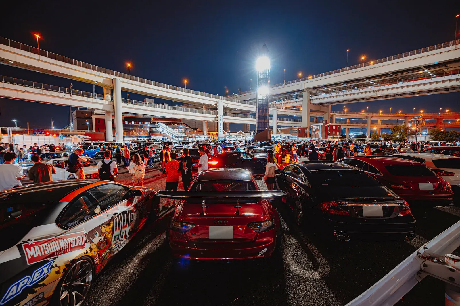 Crowd gathered around tuned cars beneath floodlit expressway ramps at night