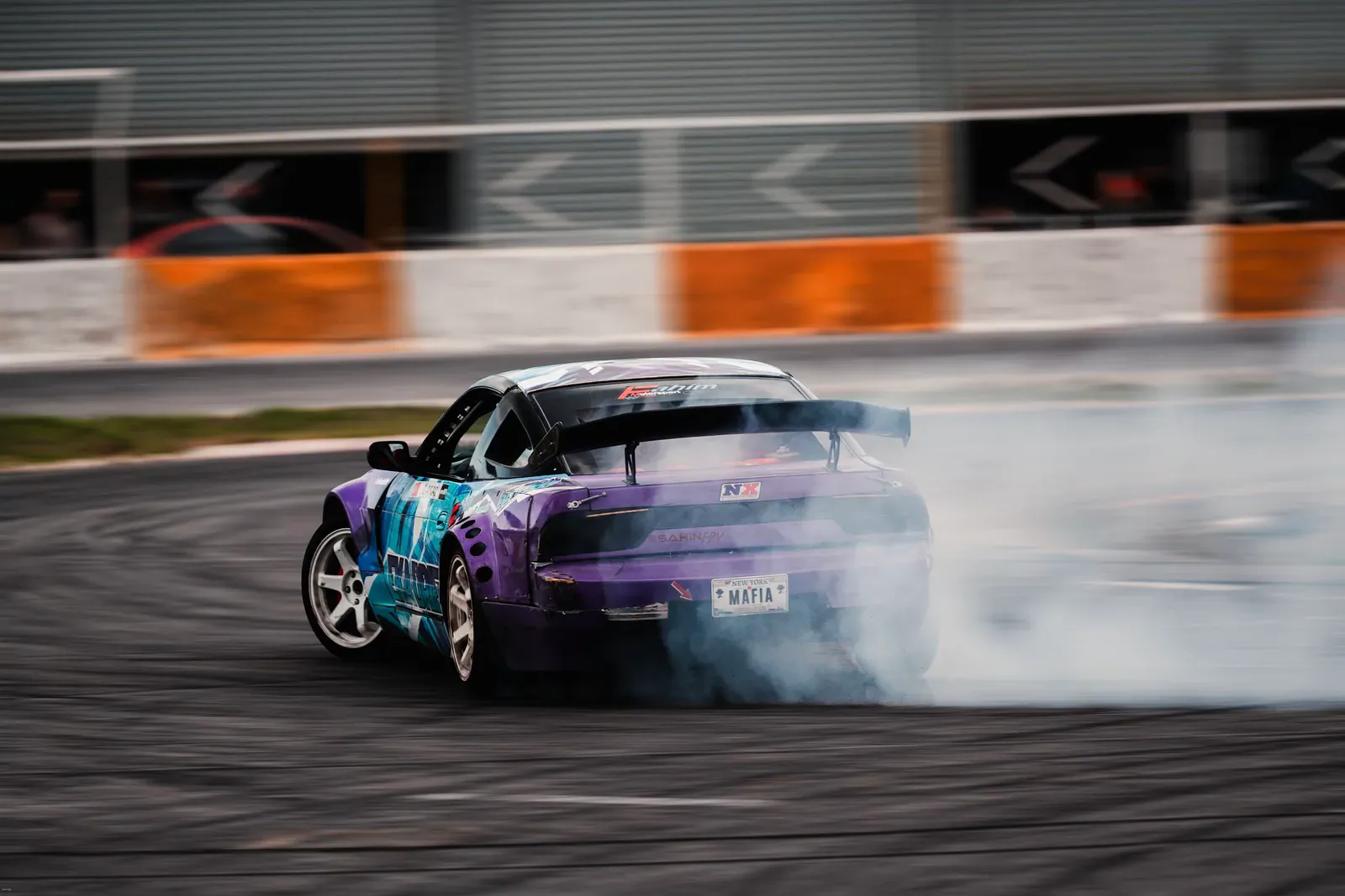 Purple and teal drift car sliding in smoke beside a pit wall