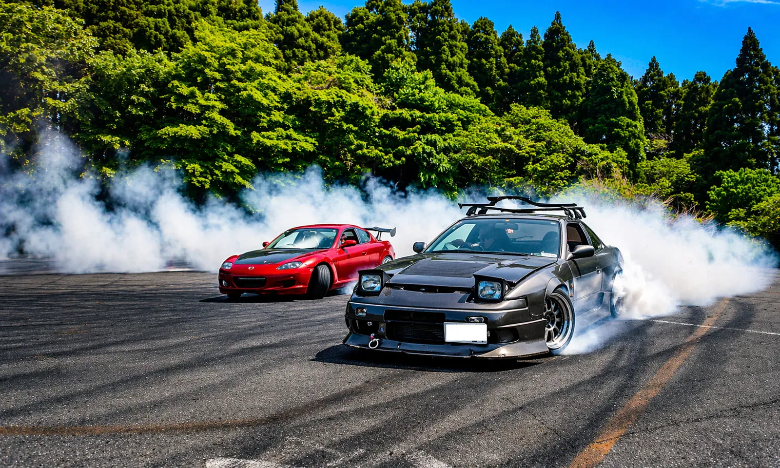 Red and black drift cars sliding through white smoke beside green forest