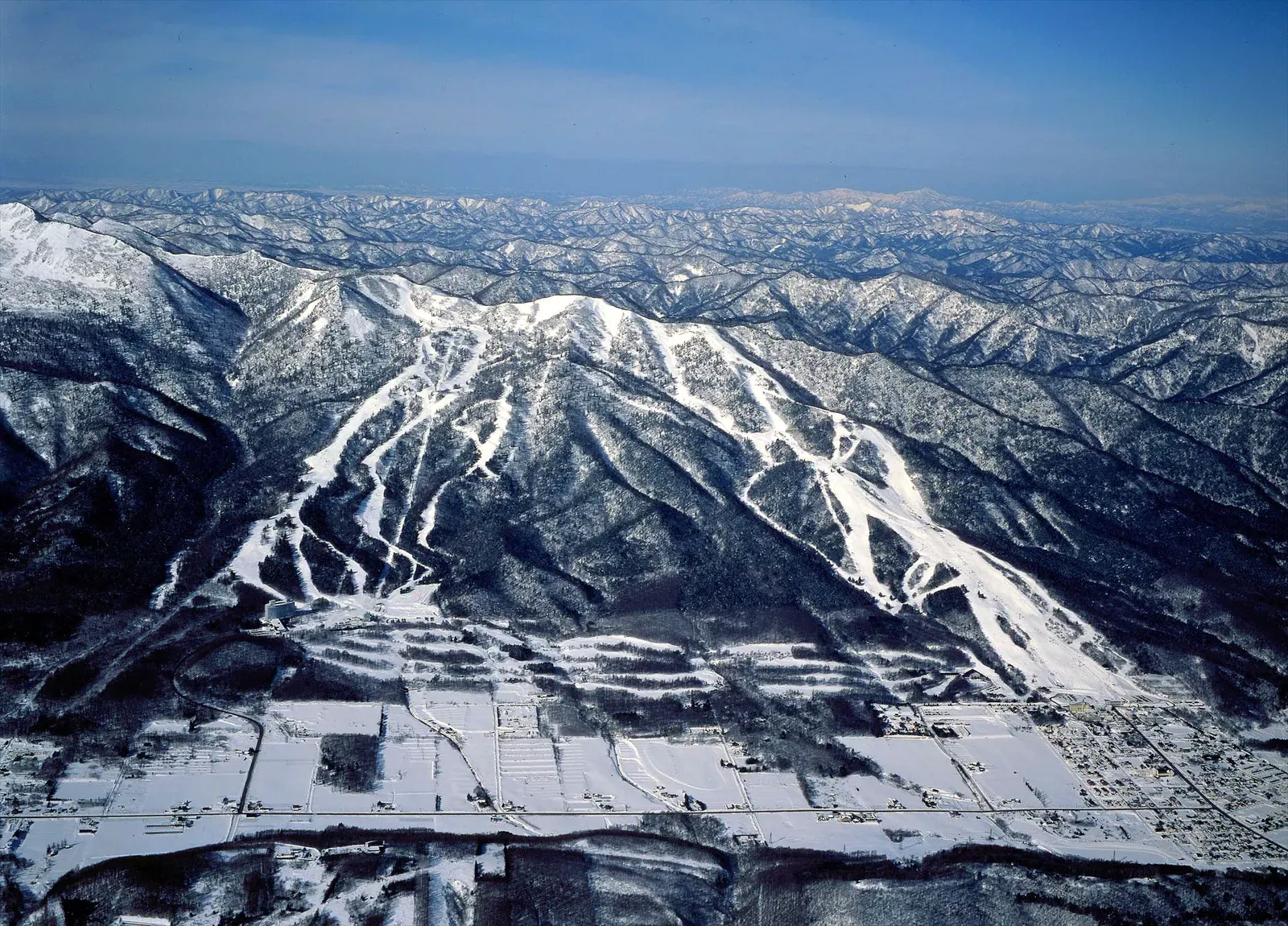 Aerial view of Furano ski runs cut through forested mountains above farmland