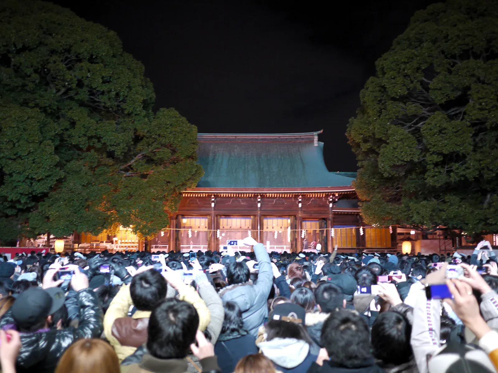 Crowd holding up phones before an illuminated shrine hall at night