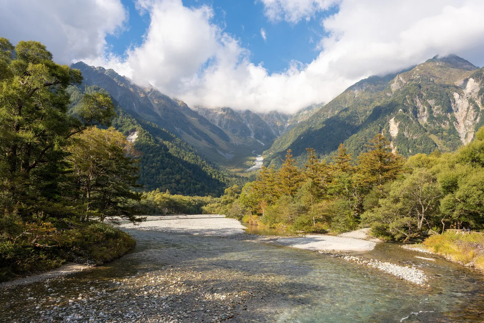 Blue river in Kamikochi valley with forested mountains and clouds beyond