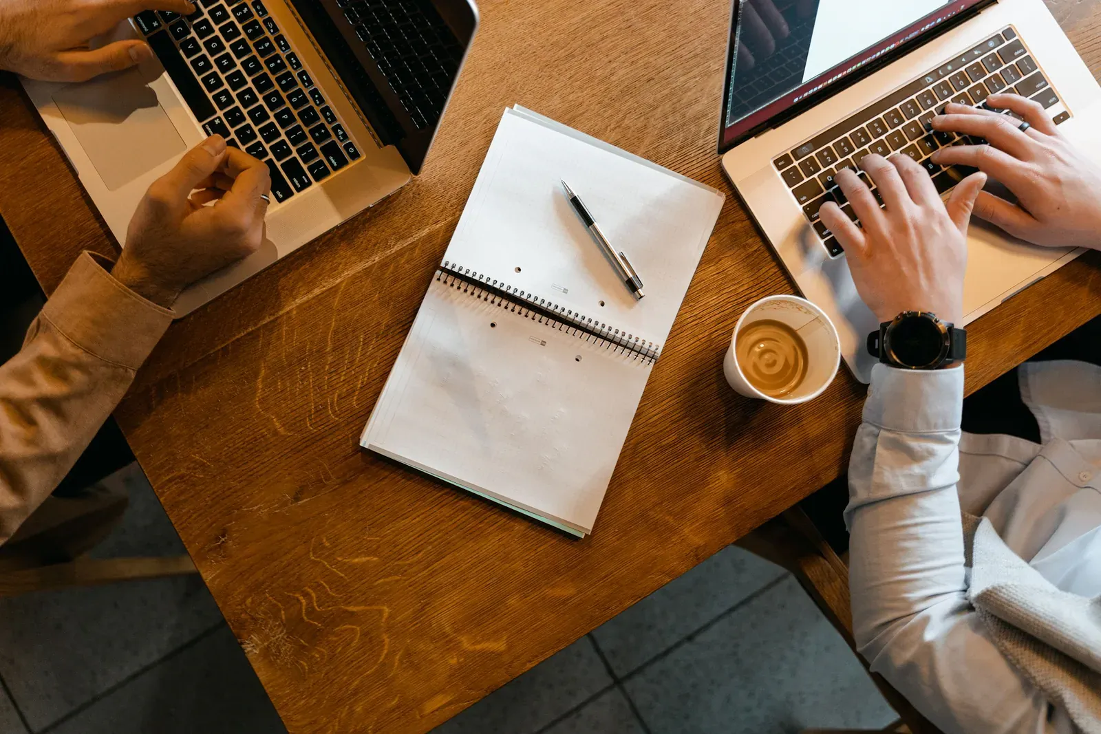 Two people typing on laptops at a wooden cafe table with notebook and coffee