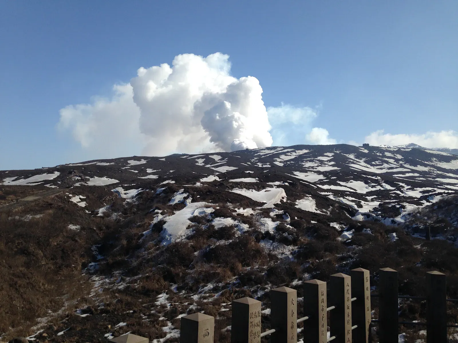 White steam plume above a snow-patched volcanic slope behind a fence