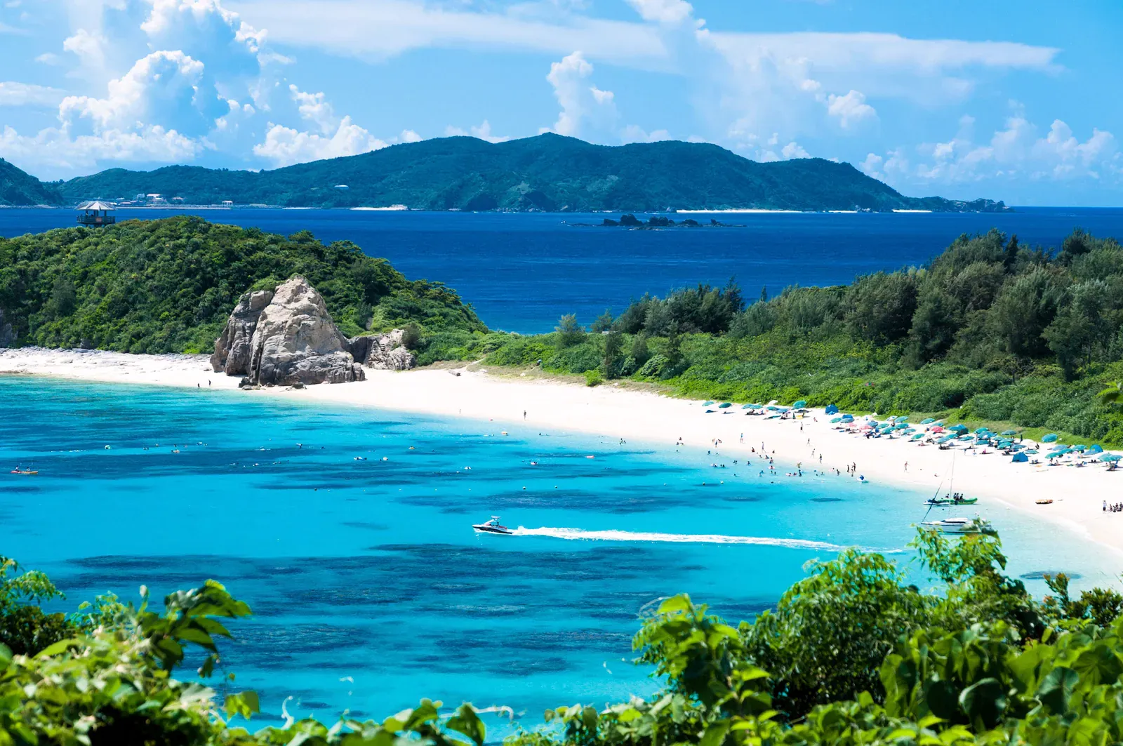Boats and swimmers on a white-sand beach in turquoise water below green hills