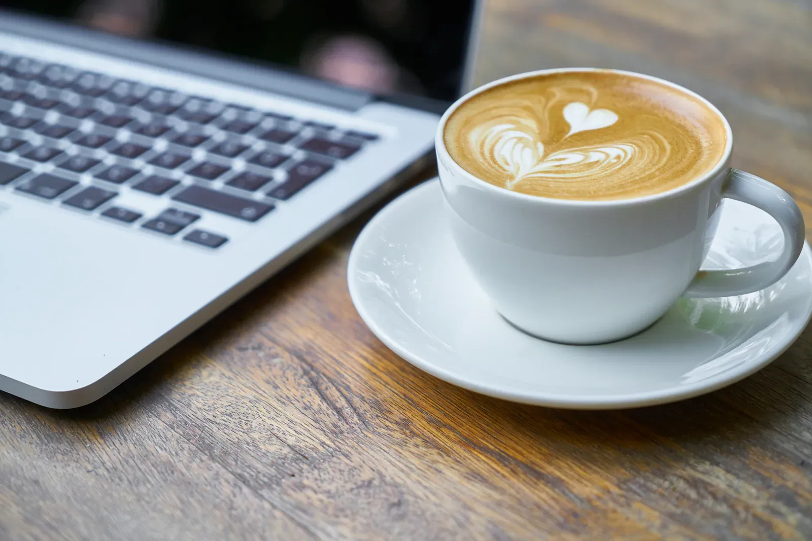 Laptop beside a latte with heart-shaped foam on a wooden cafe table